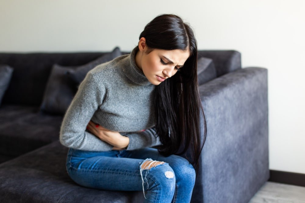 Rancho, California young woman sitting on a sofa holding her stomach in pain.