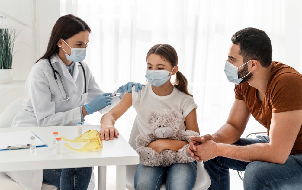 Rancho, California clinic with a female doctor giving a vaccination to a young girl holding a teddy bear, while her father watches.